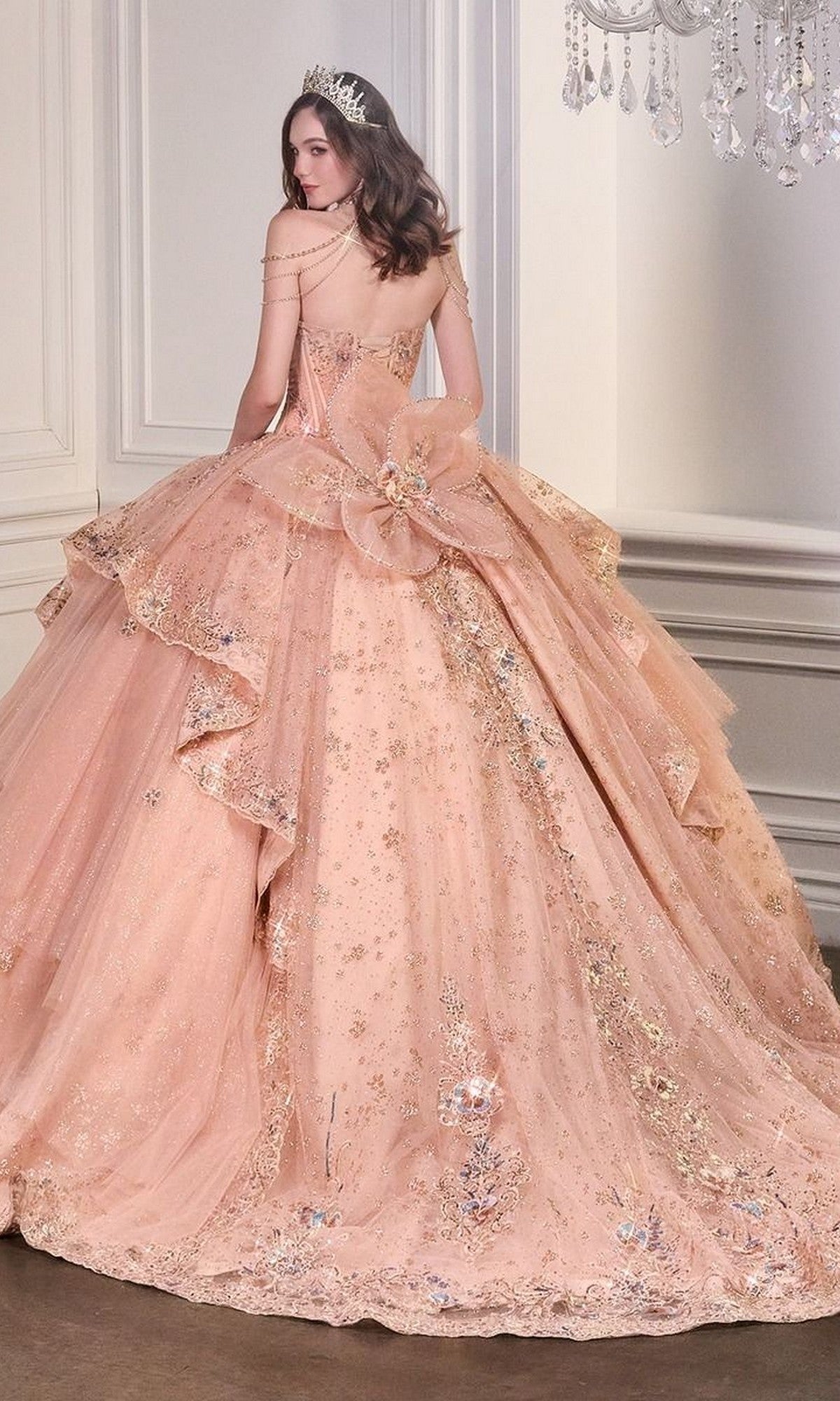 A woman wearing the Quinceanera Dress 15727 By Ladivine, featuring sparkling rose gold fabric and floral embellishments, paired with a tiara, stands in an elegant white room.